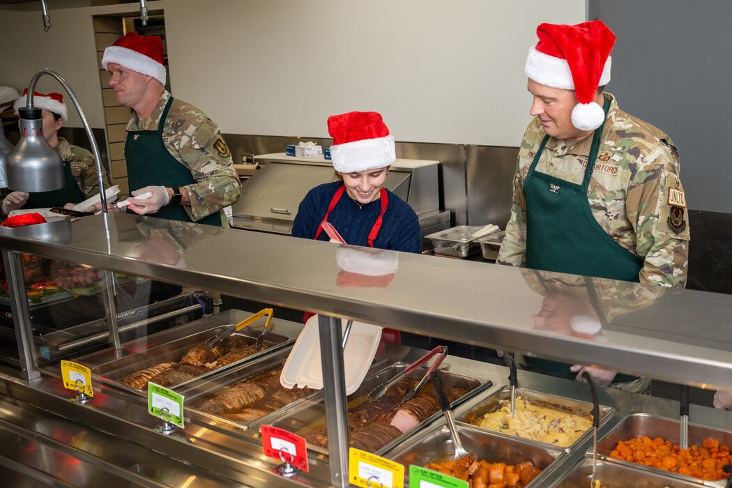 Sarah, young girl in a Santa hat, stands behind a meal prep counter passing a white Styrofoam tray to Col. Pond, in military uniform and Santa hat.