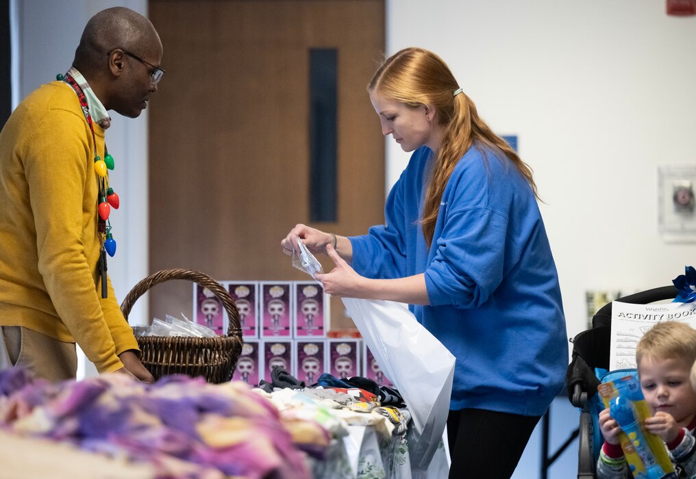 A man and woman face each other  across a table  with clothes and boxes on it. In the corner of the photo, a toddler in a stroller holds up and looks at a package.