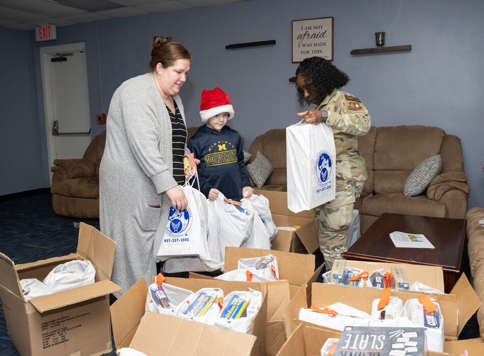 A woman in civilian clothes and a child in a Santa hat, hold bags while a woman in uniform hold another. Boxes filled with games fill the foreground.