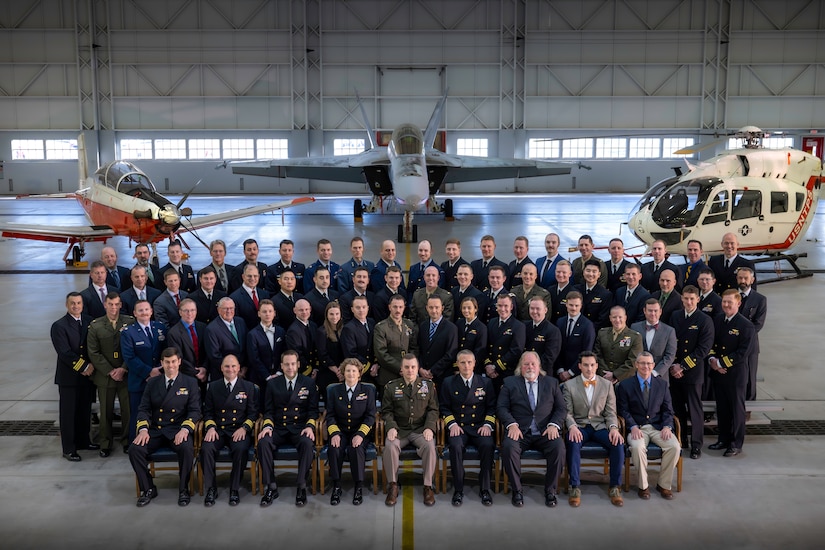 Graduating pilots pose in a group photo in front of helicopter and two aircraft.