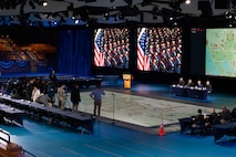 panned out shot of stage with service members at a table and media across the stage. also has screens in background displaying a map and past photo of service members