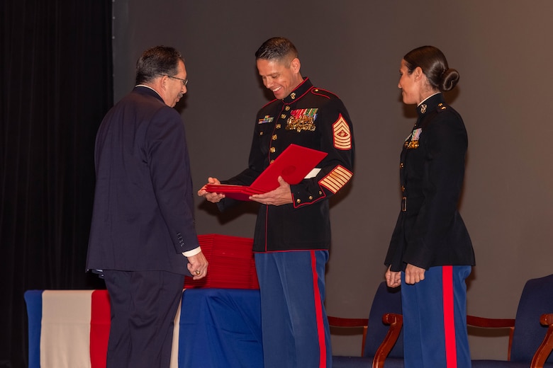 U.S. Marine Corps Sgt. Maj. Carlos A. Ruiz, Sergeant Major of the Marine Corps, center, presents a Certificate of Commendation to retired Sgt. Domenic Ferlaino a former Marine Security Guard assigned to Detachment Cameroon at Little Hall on Marine Corps Base Quantico, Virginia, Dec. 13, 2024. The former Detachment Cameroon Marines attended a Marine Security Guard graduation ceremony to receive certificates of Commendation for their heroic actions taken during 1977 while rescuing an aide employee from marauders. (U.S. Marine Corps photo by Cpl. Darien Wright)