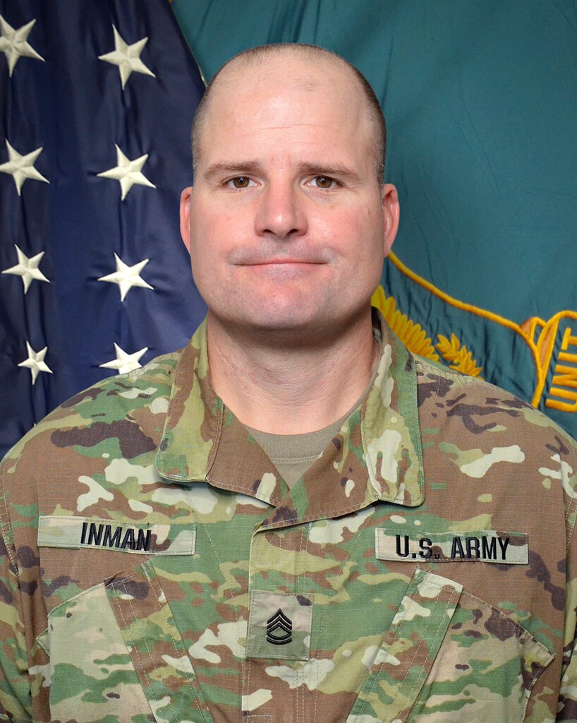 Man in U.S. Army uniform standing in front of two flags.