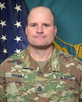 Man in U.S. Army uniform standing in front of two flags.