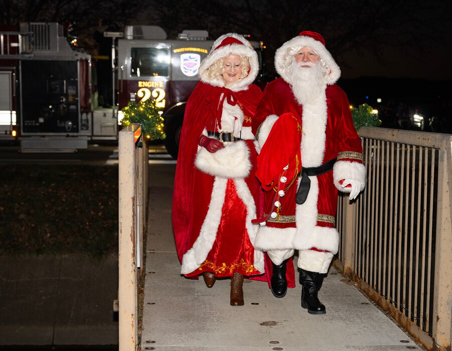Santa and Mrs. Claus cross a foot bridge at night. There is a fire truck behind them
