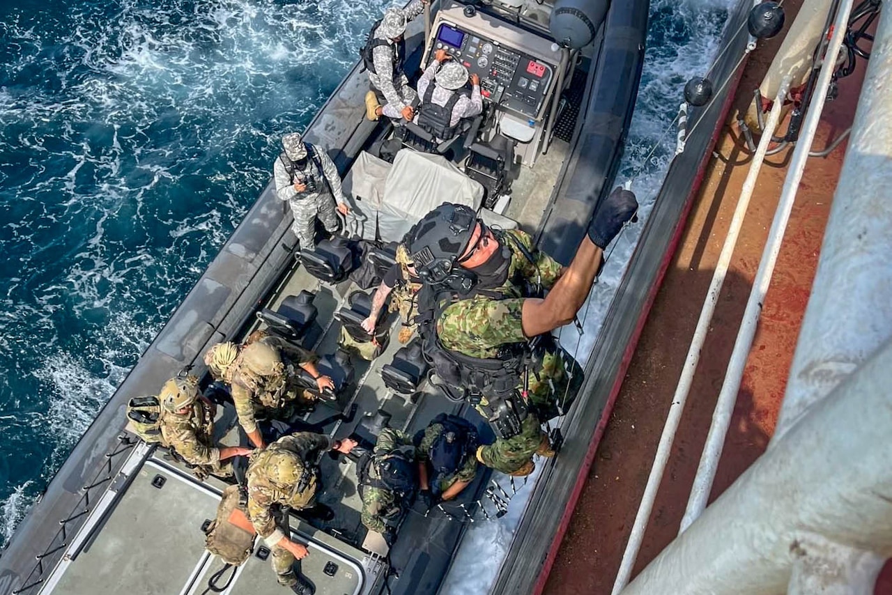 Armed sailors climb from a small boat onto a cargo ship at sea.