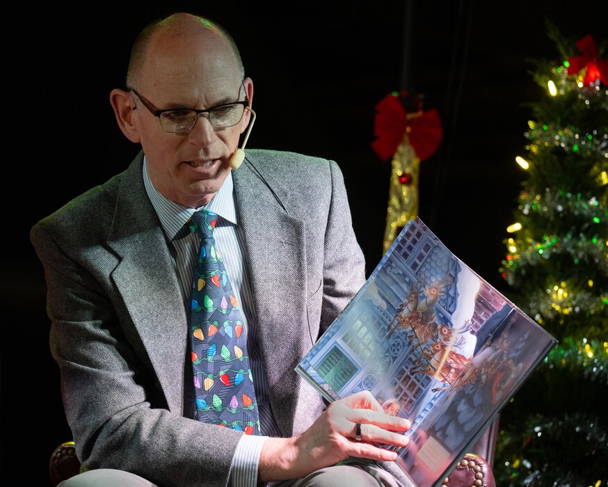 A man in a coat and tie, holds a book open as he reads aloud.