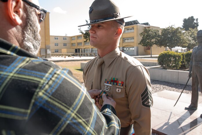 Baby Aisla’s grandpa, pins U.S. Marine Corps Drill Instructor, Staff Sgt. Barry Clayton, right, with Charlie Company, 1st Recruit Training Battalion, who is being presented with a Navy and Marine Corps Achievement Medal at Marine Corps Recruit Depot San Diego, California, Dec. 13, 2024. A NAM is given to members of the U.S. Navy and Marine Corps for meritorious service or achievement. Delacruz and Clayton received a NAM for exemplifying unrivaled courage and extraordinary presence of mind when confronting a life-threatening emergency. The words “drill instructor, drill instructor, drill instructor” were called and Delacruz and Clayton sprang into action to save a fourteen-month-old infant who was unconscious and not breathing. Providing CPR and proper throat sweeping techniques they successfully dislodged the obstruction and the infant regained consciousness. Likely saving baby Aisla’s life.  The family of the infant was onsite during the ceremony and presented the awards. The Marine Corps has served the Nation honorably for 250 years. Marines embody the uncompromising standards and characteristics of honor, courage and commitment.