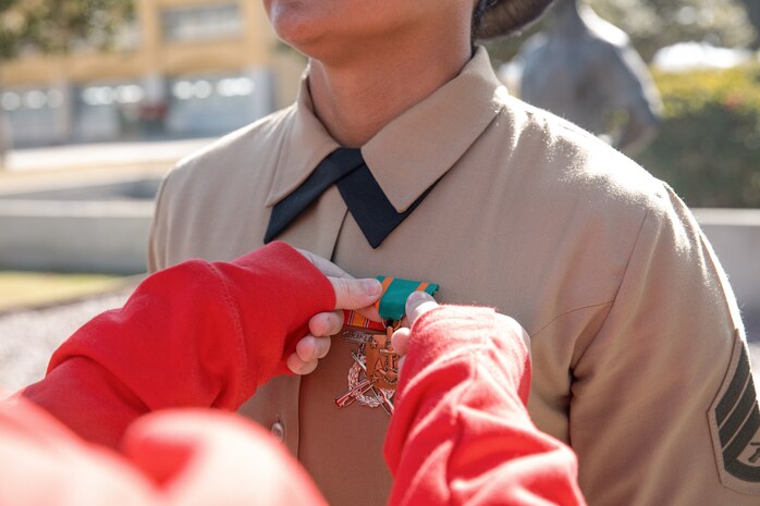 Baby Aisla’s mom, pins U.S. Marine Corps Drill Instructor, Staff Sgt. Burruel Delacruz, left, with Charlie Company, 1st Recruit Training Battalion, who is being presented with a Navy and Marine Corps Achievement Medal at Marine Corps Recruit Depot San Diego, California, Dec. 13, 2024. A NAM is given to members of the U.S. Navy and Marine Corps for meritorious service or achievement. Delacruz and Clayton received a NAM for exemplifying unrivaled courage and extraordinary presence of mind when confronting a life-threatening emergency. The words “drill instructor, drill instructor, drill instructor” were called and Delacruz and Clayton sprang into action to save a fourteen-month-old infant who was unconscious and not breathing. Providing CPR and proper throat sweeping techniques they successfully dislodged the obstruction and the infant regained consciousness. Likely saving baby Aisla’s life.  The family of the infant was onsite during the ceremony and presented the awards. The Marine Corps has served the Nation honorably for 250 years. Marines embody the uncompromising standards and characteristics of honor, courage and commitment.