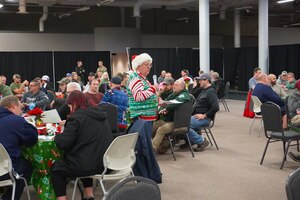 A man wearing a Santa hat and holiday sweater speaks to a group of people sitting at tables.