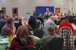 A group of people sitting at tables in a dining hall look at a man in a camouflage military uniform who is speaking into a microphone.