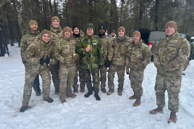 A group of guardsmen pose for a photo wearing cold-weather gear and standing on snow in a forest.