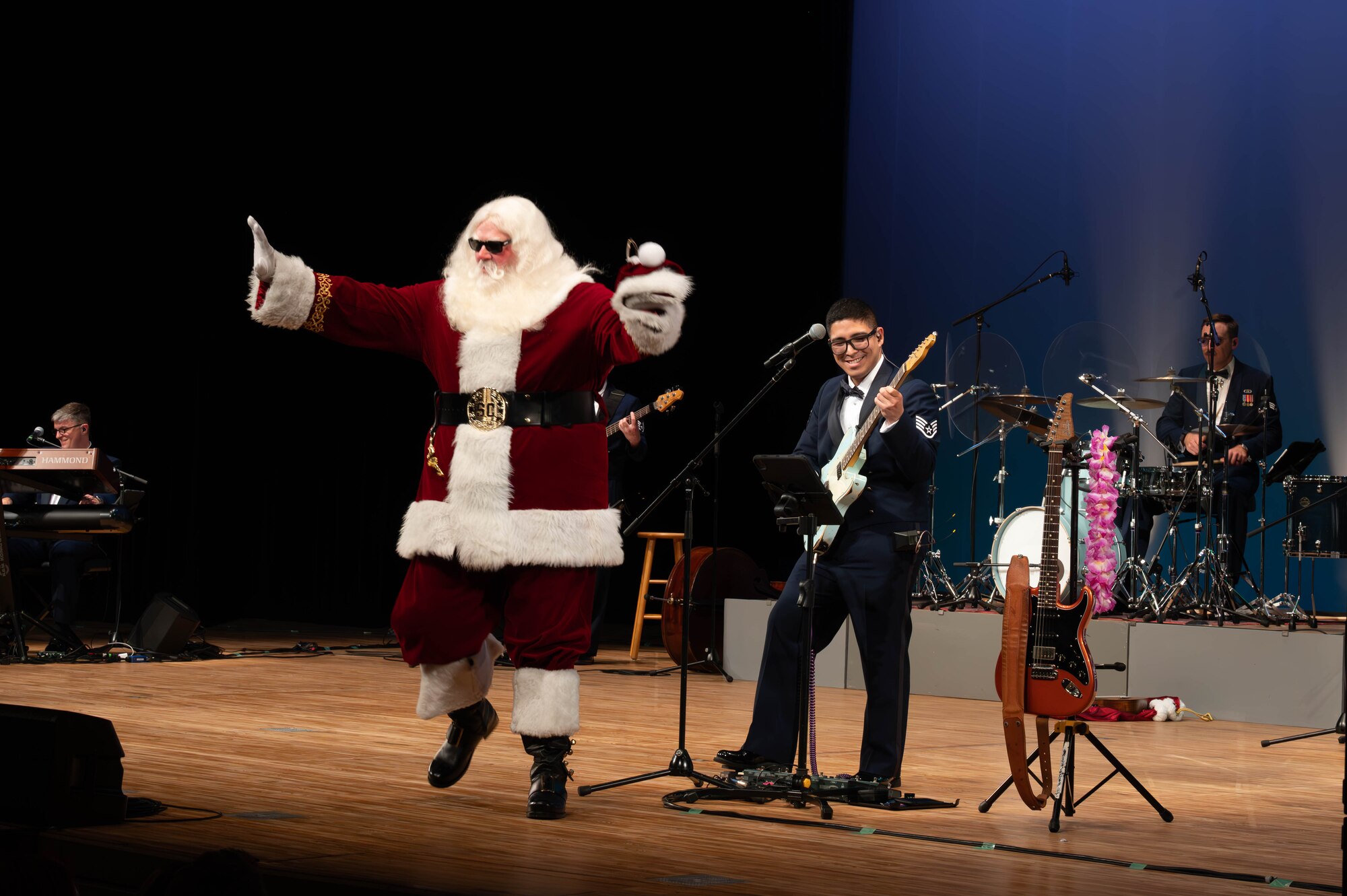 A man in a Santa uniform waves towards the crowd and a guitarist in military blues plays and smiles on stage.