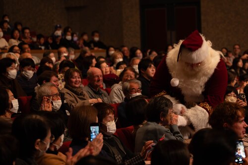 Santa,  holding an elderly lady's hand while audiences take photos of him through the crowd.
