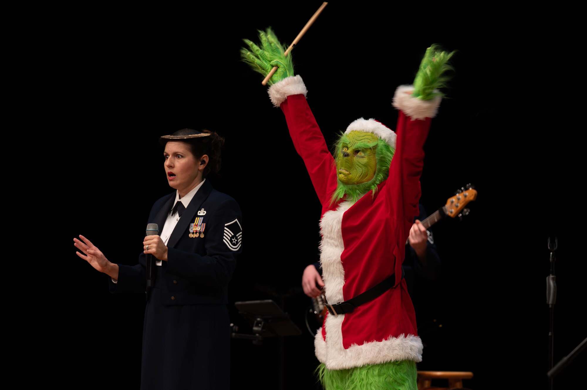 U.S Air Force military band member wearing a hi-hat cymbal with a face of shock while the the grinch wearing a Santa outfit has his arms raised in triumph .