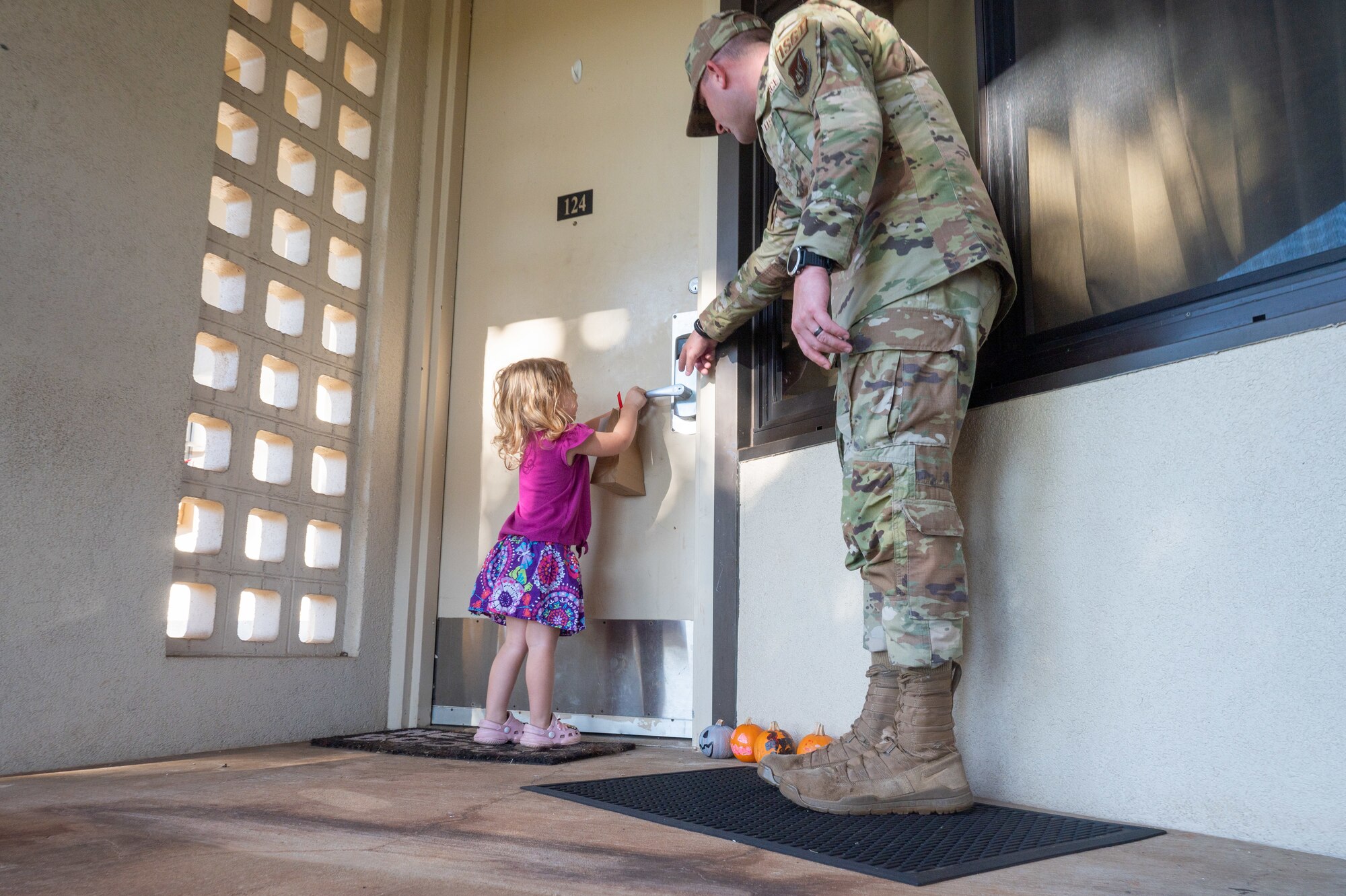 A child puts a bag full of cookies on a door handle with the help of an Airman next to her.