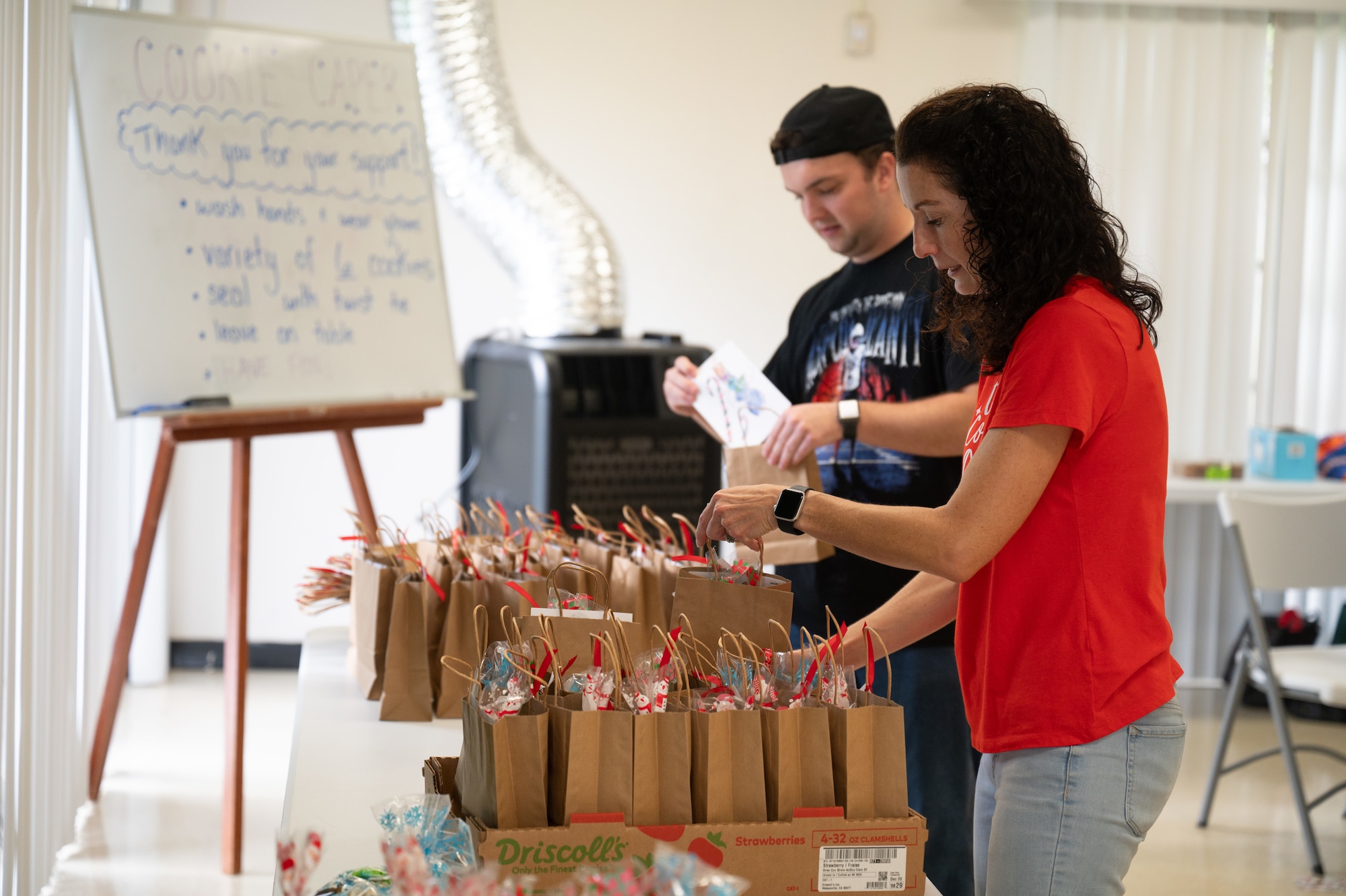Two people stand in front of a table full of bags with cookies inside of them.