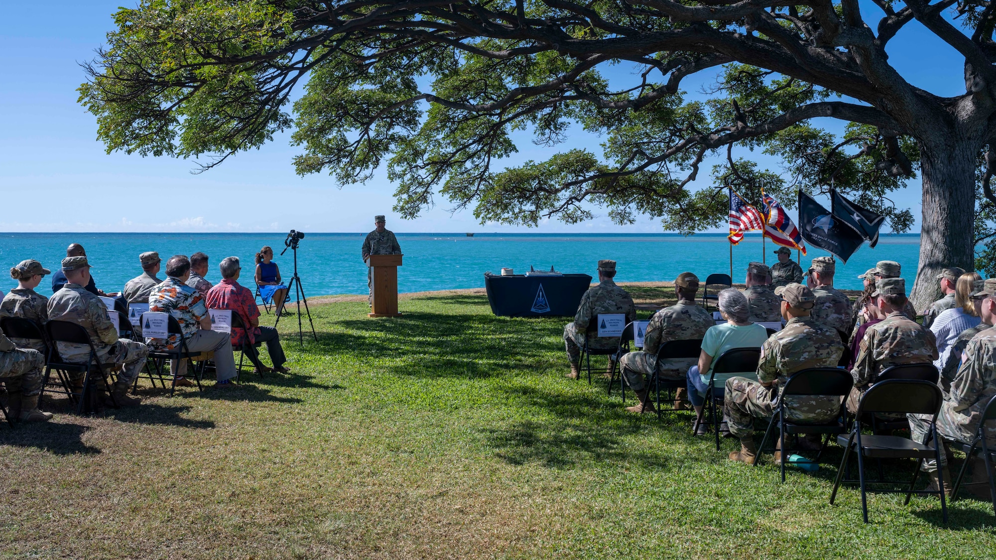 a military uniformed man gives a speech as a large, seated crowd listen