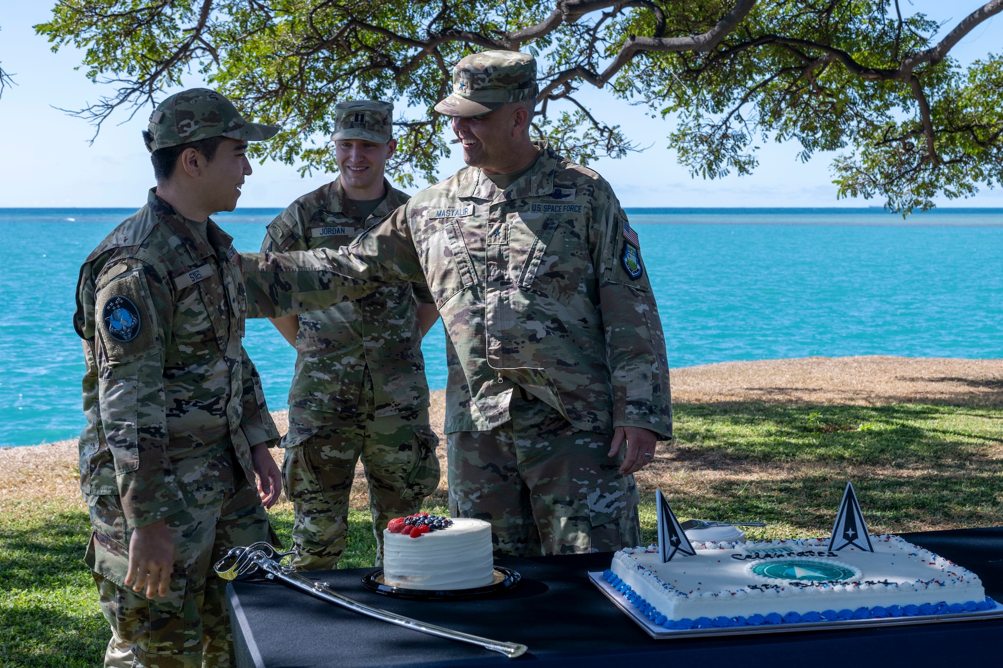 An older man in military uniform slaps a younger soldier on the shoulder as they stand in front of a table laden with a large cake