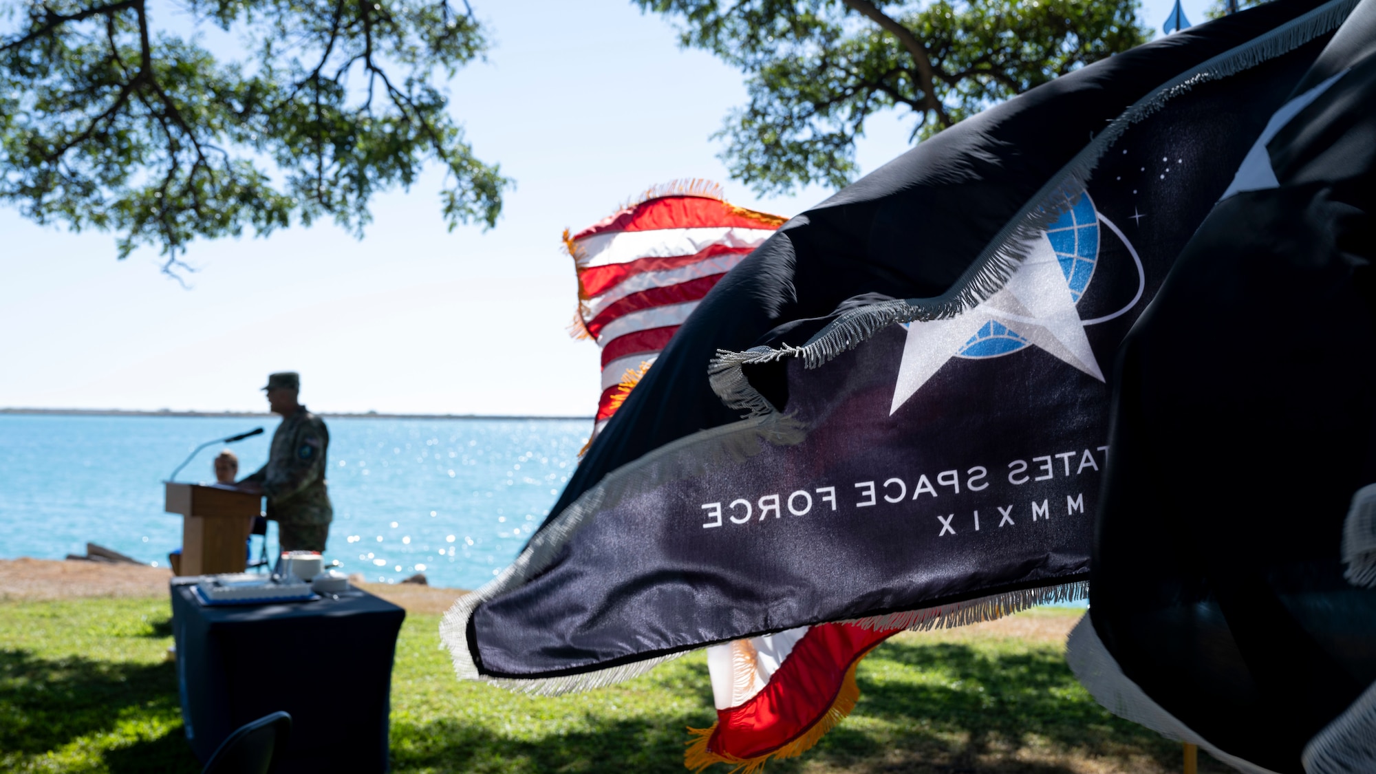 Four flags wave in the wind side by side, as a military uniformed man at a podium speaks into a microphone behind them.