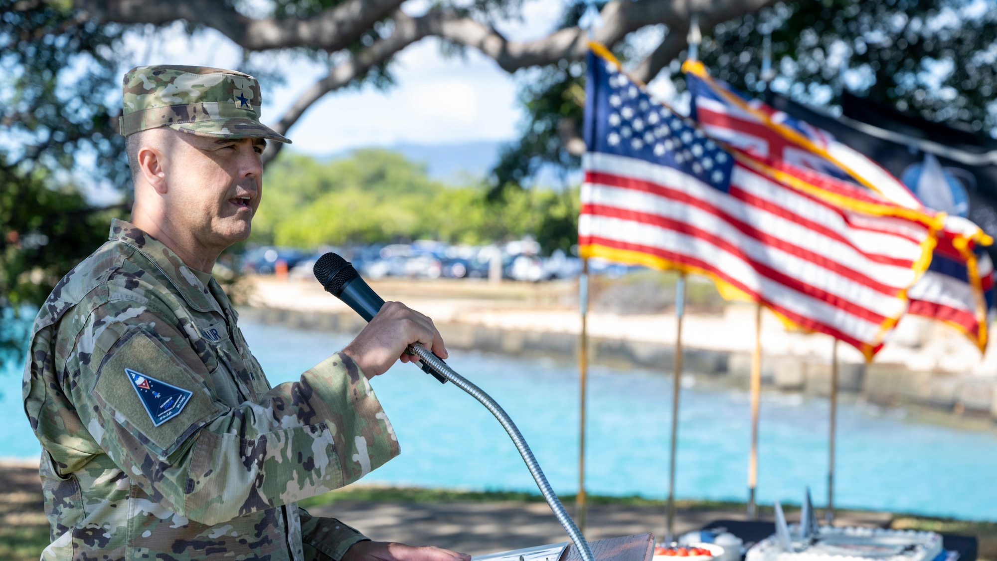 An older man in a military uniform stand at a podium with flags waving in the wind behind him.