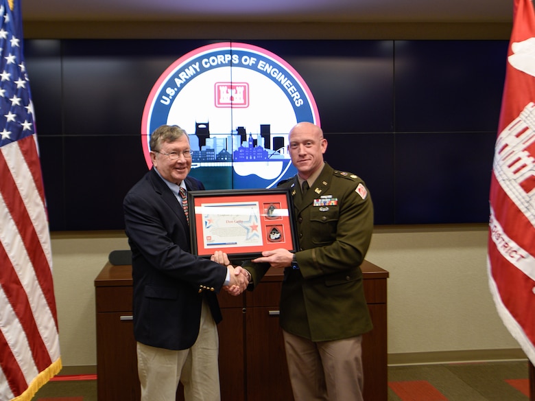 One man in a civilian suit and one man in a military uniform shake hands while holding a framed award certificate.