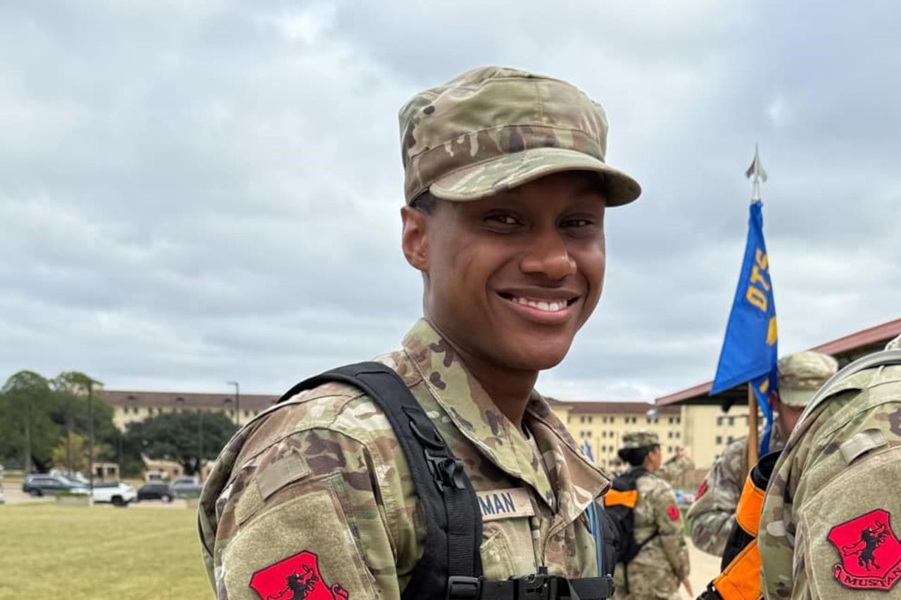 An airman poses for a photo outside by a field and some buildings during an overcast day.