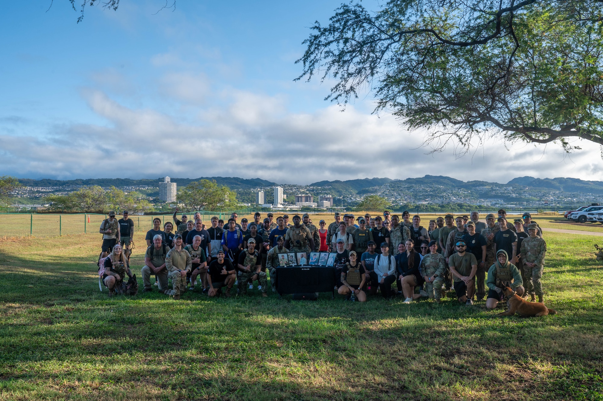 Military personnel stand together for a group photo in a green field with mountains in the background.