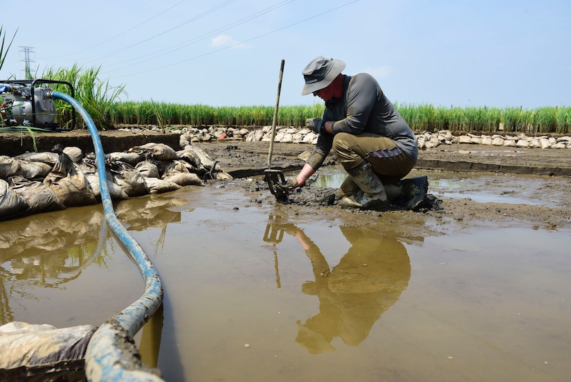 An airman kneels and holds soil with one hand in a wet, muddy area.