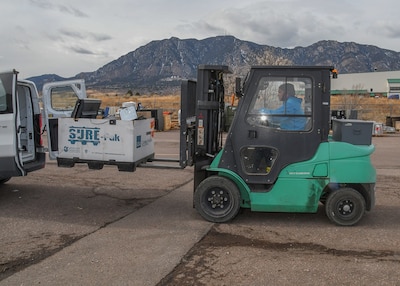 A person sits inside the cab of a green forklift. they are using it to remove property from the back of a white van. This is taking place outside and there are mountains in the background.