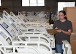 A woman holds a clipboard as she writes on it. she is standing in a Wearhouse looking towards hospital beds.