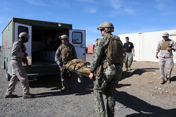 Capt. William McGinnis and Chief Hospital Corpsman (HMC) David Potter load a high-fidelity simulated manikin into an ambulance after receiving care from Expeditionary Resuscitative Surgical System (ERSS) team while Hospital Corpsman Second Class (HM2) Kwame Obengyeboah provides security. McGinnis and Potter are the two personnel that make up the En-Route Care System (ERCS) Team 48 that provides care to patients during transportation from one echelon to another. Both McGinnis and Potter are in the Reserves sharpening their skills while fulfilling the roles of a Critical Care Nurse and Search and Rescue Medical Technician, respectively. Obengyeboah is an instructor at NEMWDC and plays the role of Medical Quick Reaction Force (MEDQRF) during the ERSS course guiding the BRC students in Role 1 care.