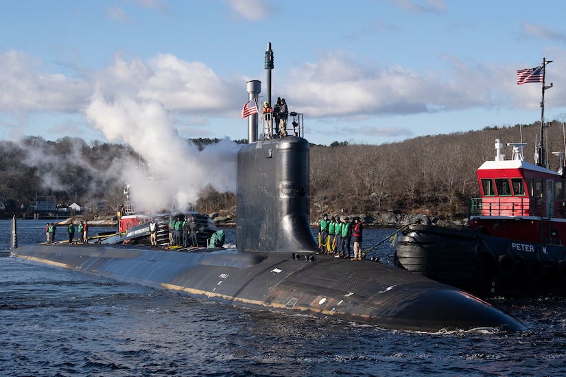 Sailors stand atop a submarine near a tugboat off a shoreline.