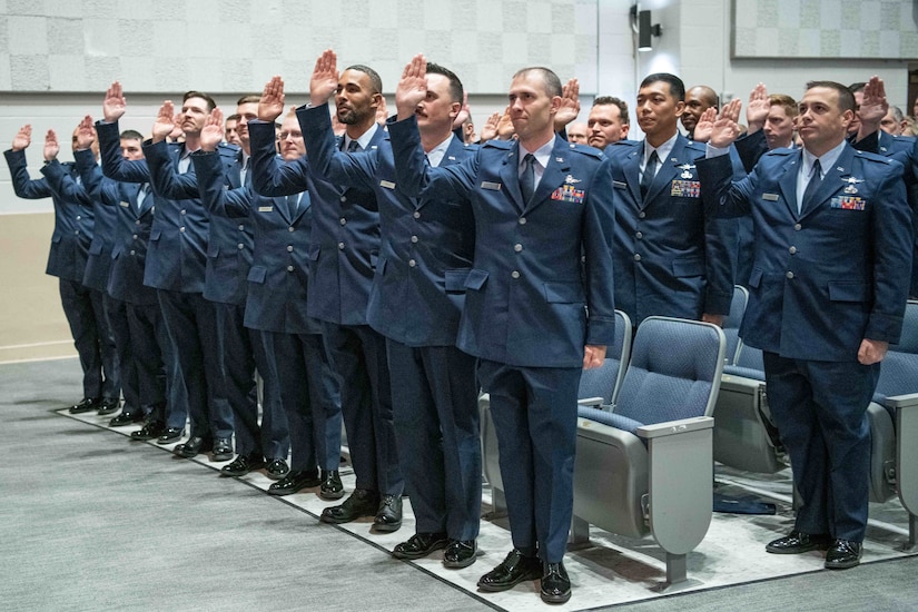 Airmen in a classroom stand with right hands raised.