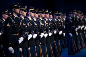 Dozens of soldiers in ceremonial uniform stand in formation in a dark room during a ceremony.