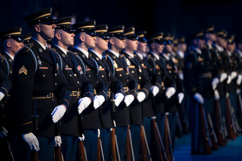 Dozens of soldiers in ceremonial uniform stand in formation in a dark room during a ceremony.