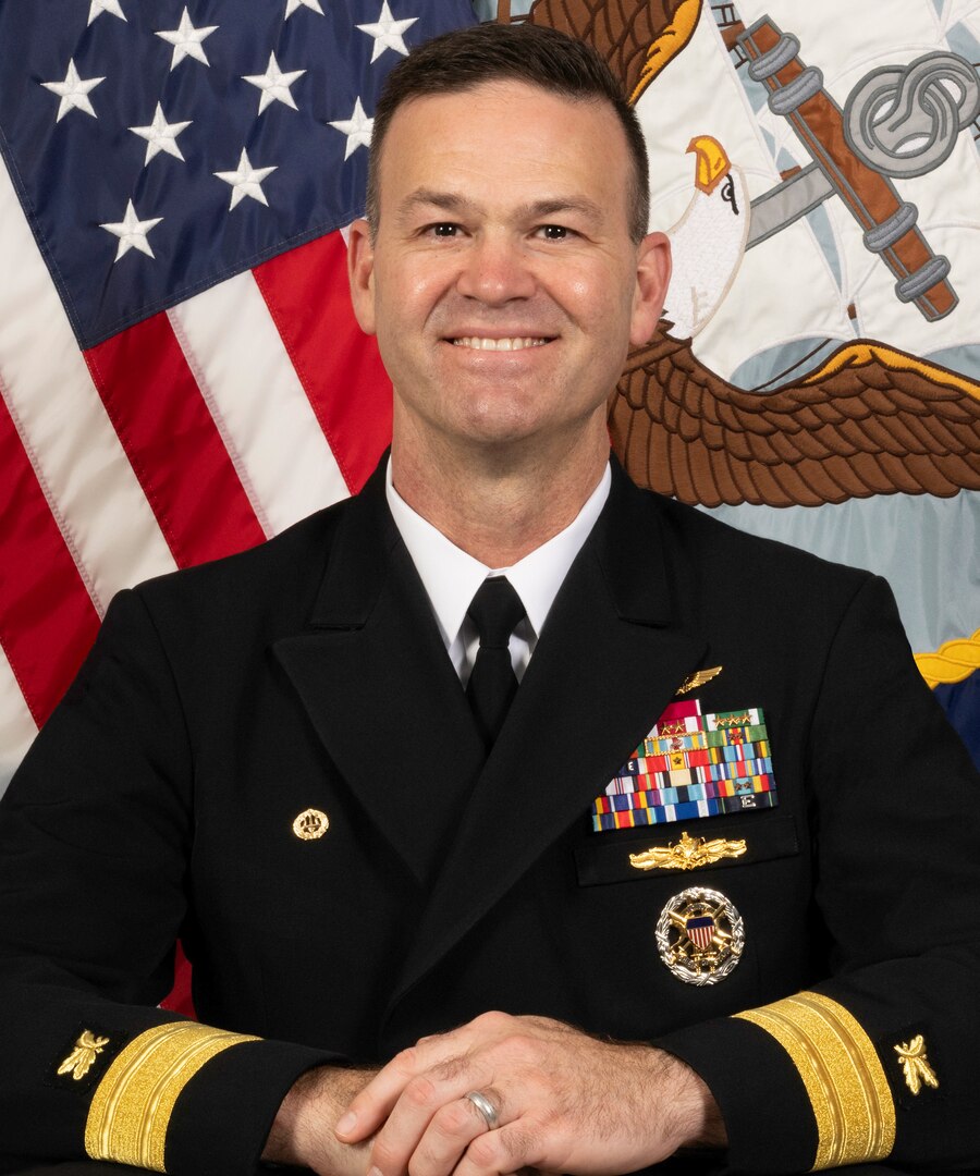 A man dressed in a U.S. Navy uniform poses for a portrait in front of flags.