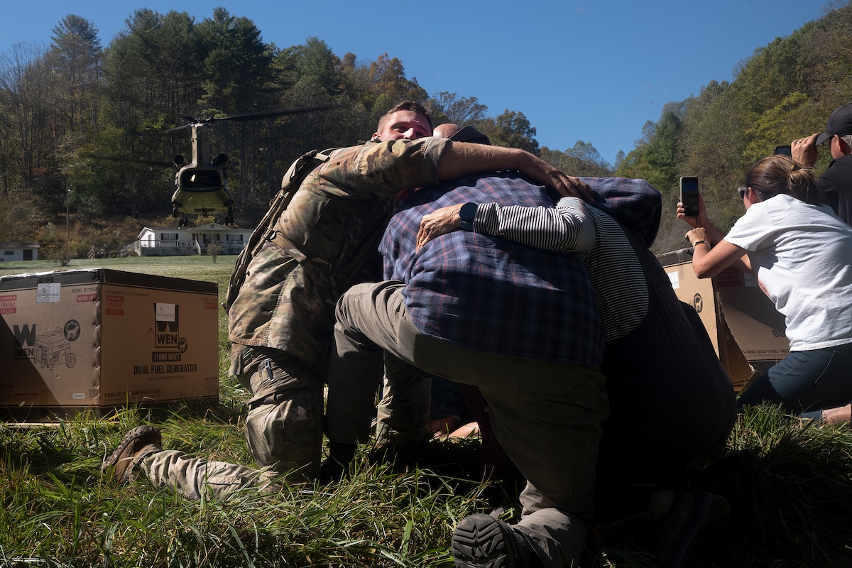 U.S. Army Sgt. Isaac Bradshaw, a combat medic with C Company, 230th Brigade Support Battalion, North Carolina Army National Guard, wraps his arms around an older couple and uses his body to block them from debris and strong winds produced by the rotor wash of a landing CH-47 Chinook helicopter in Burnsville, North Carolina, Oct. 10, 2024. The Chinook crew responded in the aftermath of Hurricane Helene, bringing generators, fuel, and other supplies to the remote area that was isolated when floodwaters washed away the main road in and out of the area.
