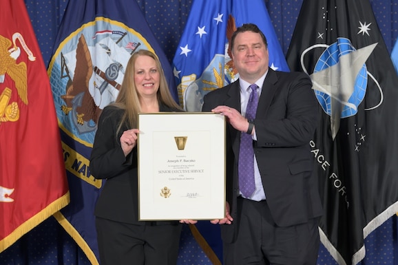 Two people stand and hold a large certificate in front of many flags.