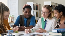 Four college students sitting in front of a computer in an office space analyzing papers in front of them and discussing their work