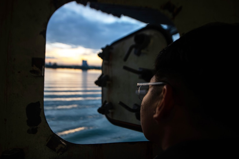 A sailor looks out a window to land from an aircraft carrier at sea.