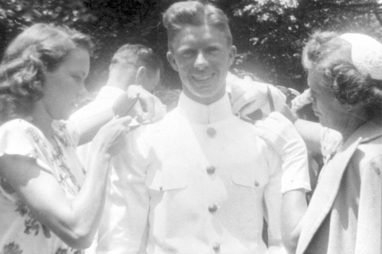 A sailor smiles as two women affix rank to his uniform.