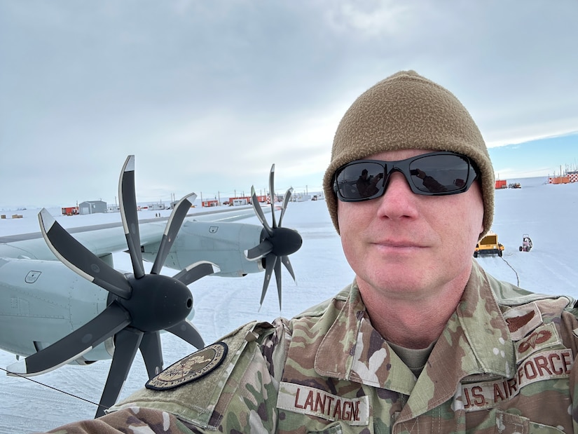 An airman wearing a winter hat, sunglasses and a camouflage uniform, stands in front of an aircraft on snow.