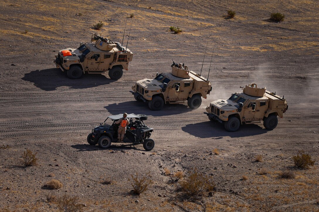 Three military vehicles drive along desert-like terrain as a person stands and watches from a smaller vehicle.
