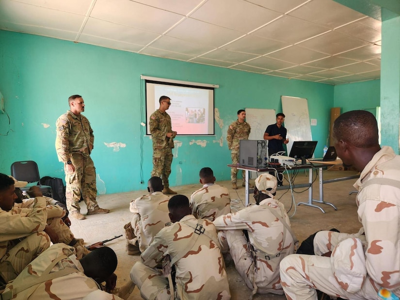 Several U.S. soldiers stand in the front of a room with light greenish walls and address seated Mauritanian troops.