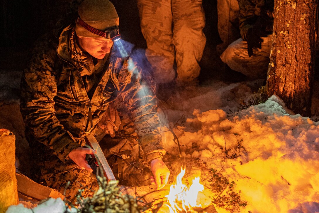 A Marine wearing a headlamp kneels by a small fire outdoors in the dark with others in the background.