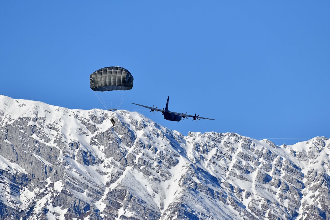 A soldier conducts a parachute jump from a large aircraft with snowy mountains in the background.