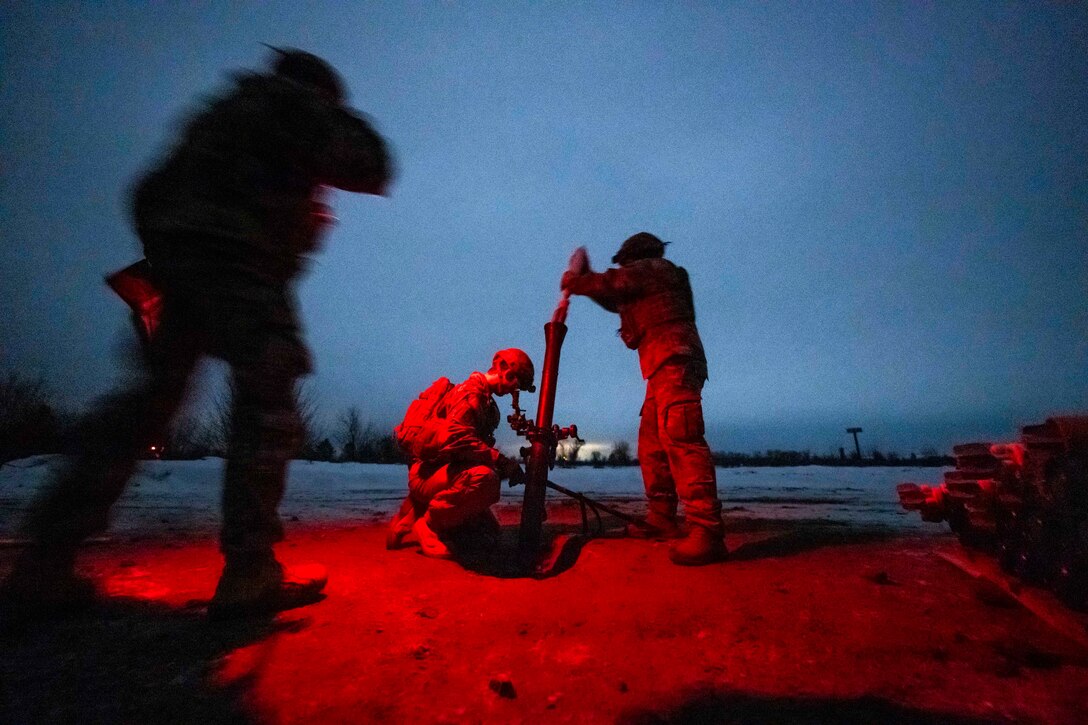 Soldiers hang training rounds on a mortar system. The three service members are illuminated by a red light.