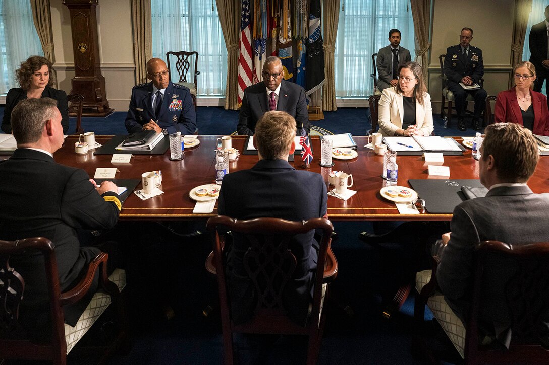 Secretary of Defense Lloyd J. Austin III and British Defense Secretary Grant Shapps sit across from each other at a large conference table with several other participants and others standing in the background.