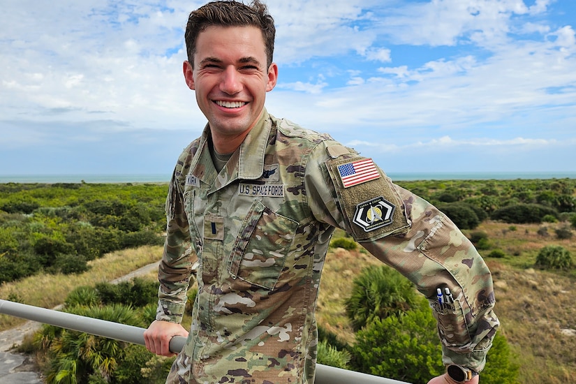 A smiling person in a military uniform poses in front of a field.